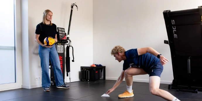 Twee sporters trainen; één knielt tijdens een oefening en de ander houdt een bal vast.