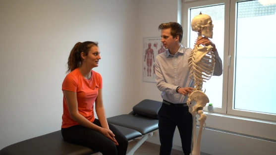 Een zorgverlener bespreekt een anatomisch skelet met een vrouw in een behandelkamer.