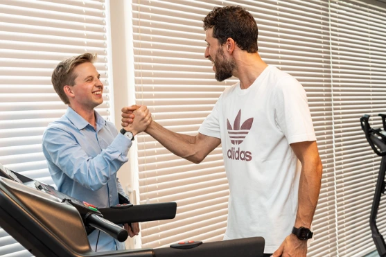 Twee lachende mannen geven elkaar een hand naast fitnessapparatuur.