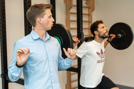 Twee mannen trainen met een halter; één voert een squat uit terwijl de ander toekijkt.