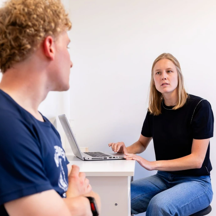 Vrouw zit aan laptop en praat met een man in een lichte kantoorruimte.